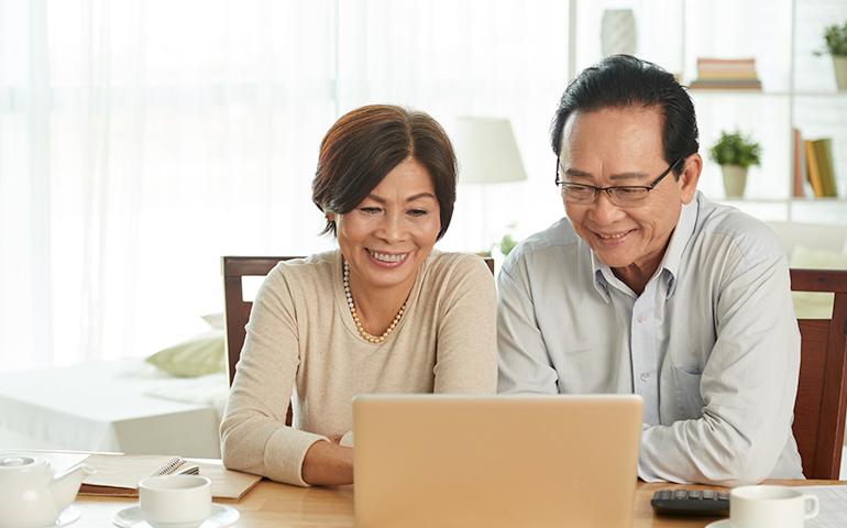 Older Asian couple sitting at home in front of a laptop. 