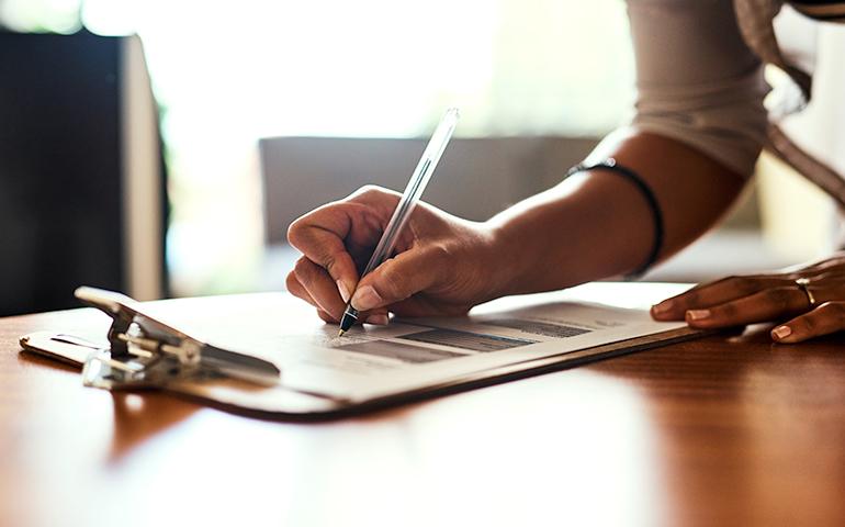 Photo of woman's hand leaning down and writing on a clip board.