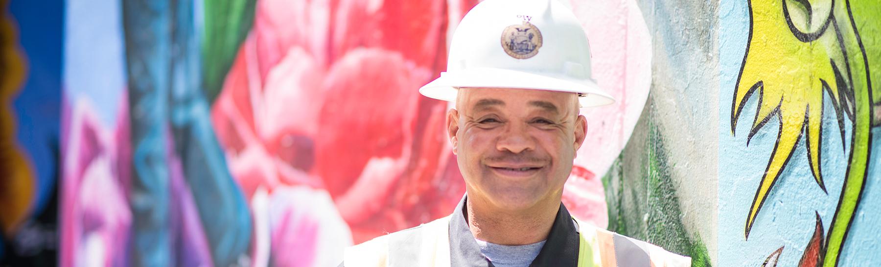 Image of older male City and County of San Francisco employee from Department of Public Works (DPW) wearing a hardhat and standing in front of the Hemlock Street Mural in San Francisco.